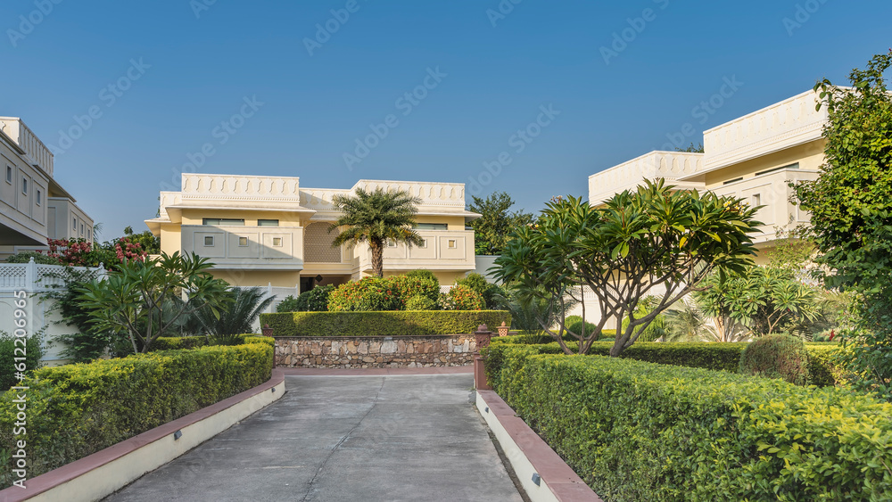 A concrete road with a hedge on the roadsides goes forward. Ahead is a decorative flower bed with trimmed and flowering bushes, a palm tree. Beige cottages against the blue sky. India.