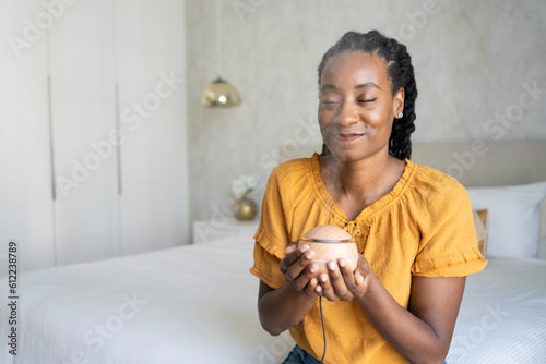 Smiling woman smelling aroma from air diffuser sitting on bed at home photo