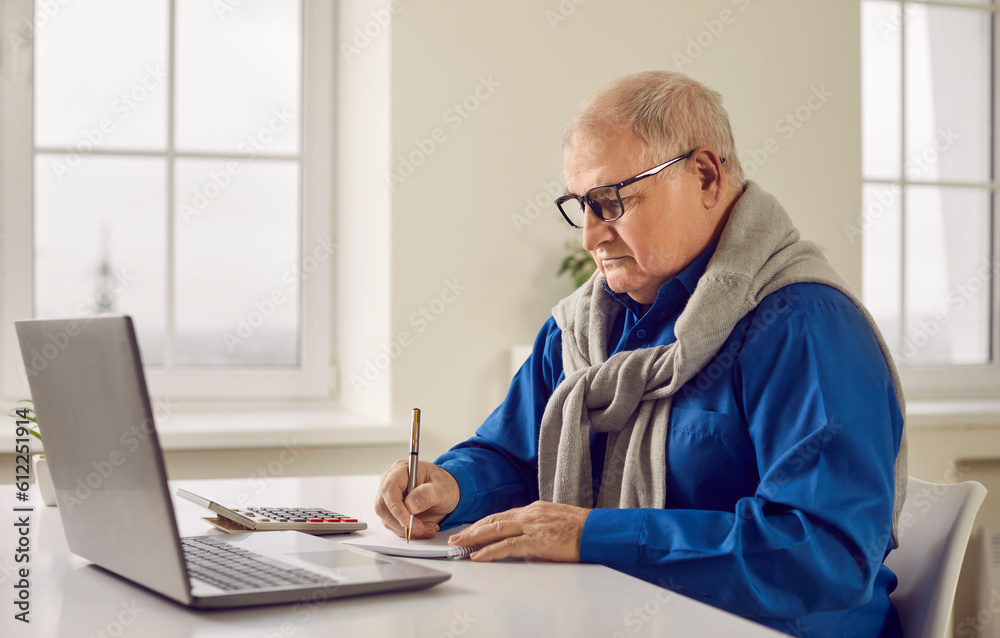 Senior retired old man pensioner in glasses sitting at desk, using modern laptop computer, working with finances, doing his accounts, planning budget, and writing something in paper notebook Senior retired old man pensioner in glasses sitting at desk, using modern laptop computer, working with finances, doing his accounts, planning budget, and writing something in paper notebook