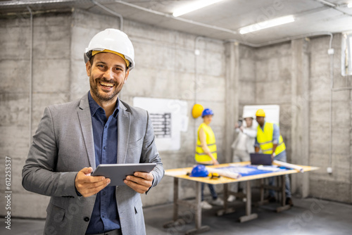 Portrait of an experienced architect with hard hat holding digital tablet computer while civil engineers planning in background.