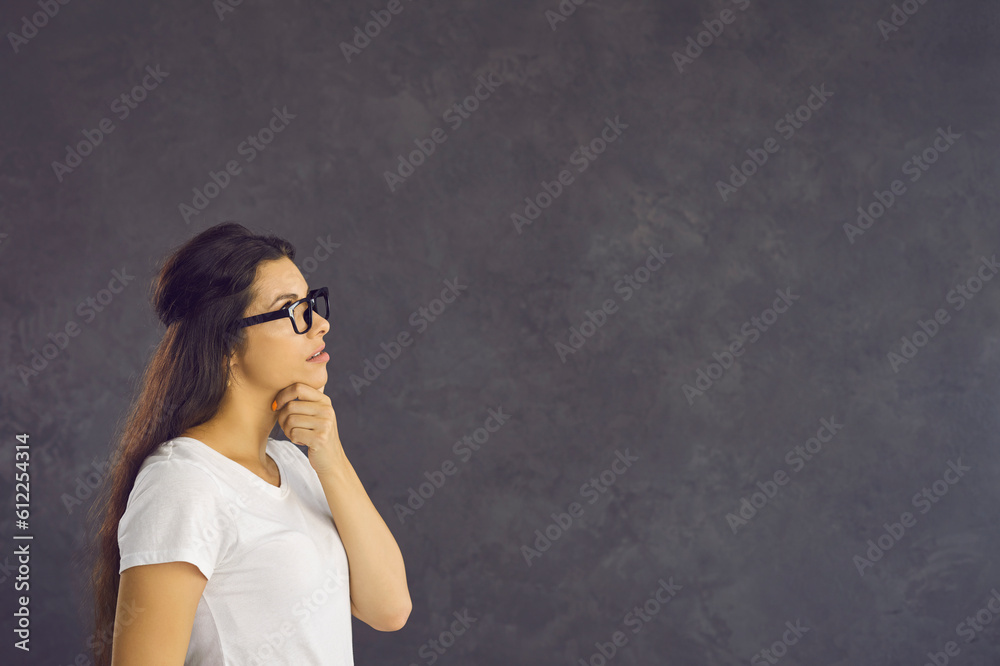 Young woman with a pensive expression stands on a gray background and thinks about something. Profile view of woman in glasses holding her chin looking at free space for text. Banner. Young woman with a pensive expression stands on a gray background and thinks about something. Profile view of woman in glasses holding her chin looking at free space for text. Banner.