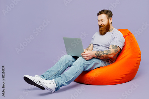 Full body young redhead bearded man wear violet t-shirt casual clothes sit in bag chair hold use work on laptop pc computer isolated on plain pastel light purple background studio Lifestyle concept