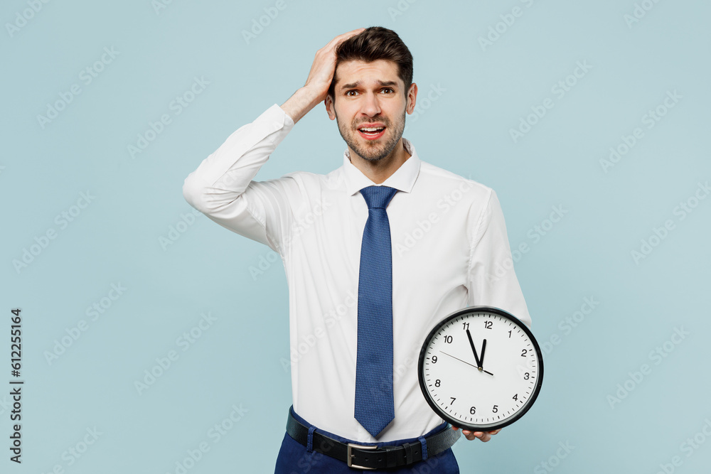 Young shocked sad troubled employee IT business man corporate lawyer wear classic formal shirt tie work in office hold head show clock isolated on plain pastel light blue background studio portrait. Young shocked sad troubled employee IT business man corporate lawyer wear classic formal shirt tie work in office hold head show clock isolated on plain pastel light blue background studio portrait.