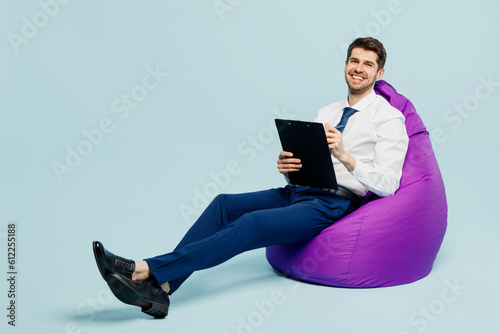 Full body happy young employee IT business man corporate lawyer wear classic formal shirt tie work in office sit in bag chair clipboard with paper account documents isolated on plain blue background.
