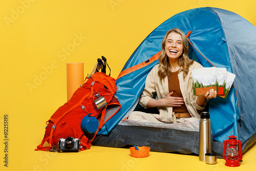 Full body young woman sit near bag with stuff tent hold tummy freeze-dried food isolated on plain yellow background. Tourist lead active lifestyle walk spare time. Hiking trek rest travel trip concept