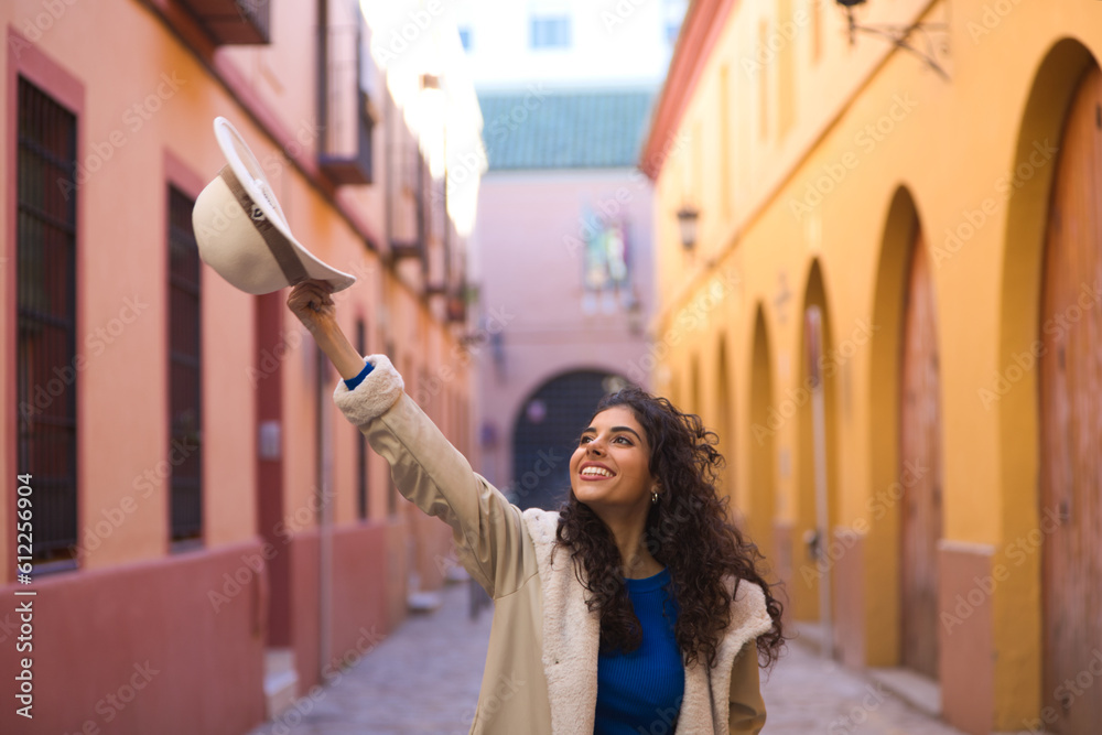 Young and beautiful Hispanic brunette woman with curly hair holds up a hat with her hand while making different expressions and having fun on her city trip in Europe.