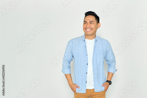 Smiling young Asian man in casual clothes posing with hands in pockets and confidently looking at copy space isolated on white background photo