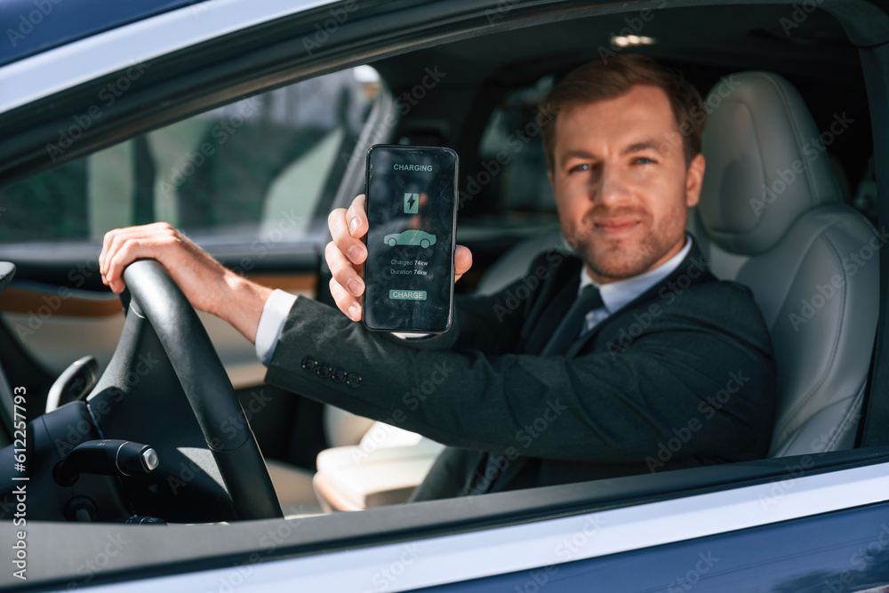 Holding smartphone with charging progress app. Businessman in suit is driving his electric car