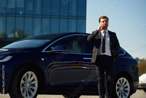 Conversation by phone. Business building behind. Man in suit and tie is standing near his car outdoors