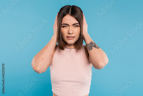 migraine, tattooed young woman with nose piercing and short hair touching head while suffering from headache on blue background, generation z, tiredness, stress