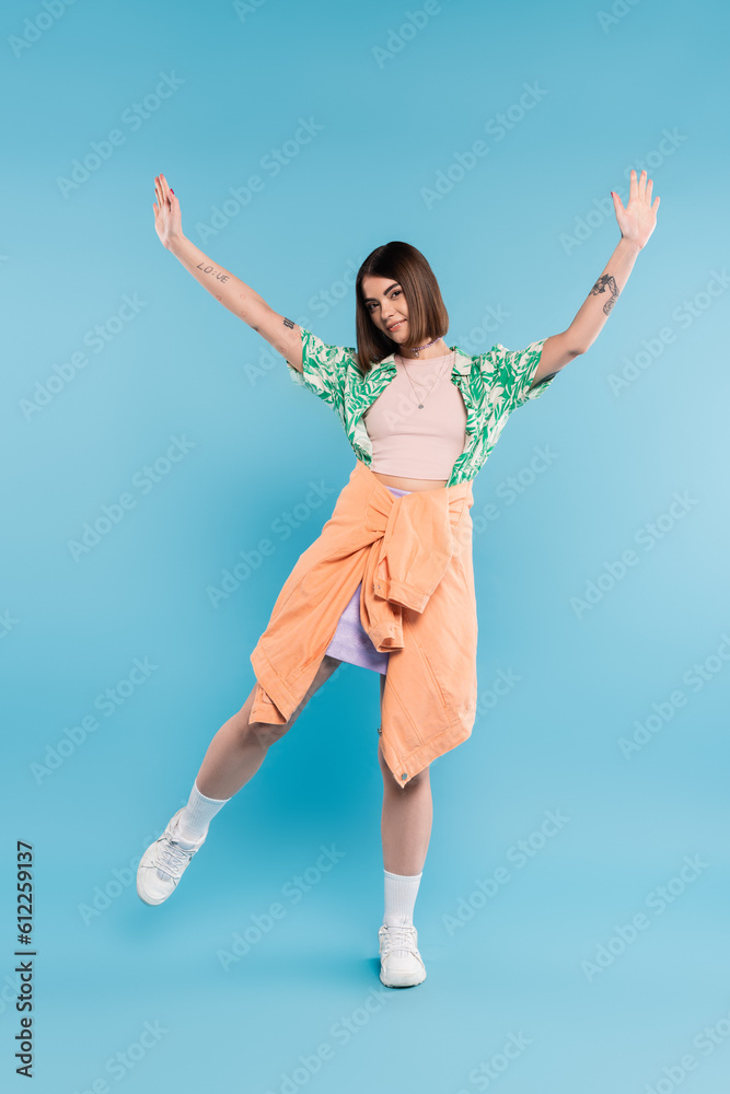 gen z fashion, carefree woman with short hair posing in shirt with palm tree print, skirt and white sneakers on blue background, tattooed, nose piercing, casual attire, raised hands, full length gen z fashion, carefree woman with short hair posing in shirt with palm tree print, skirt and white sneakers on blue background, tattooed, nose piercing, casual attire, raised hands, full length
