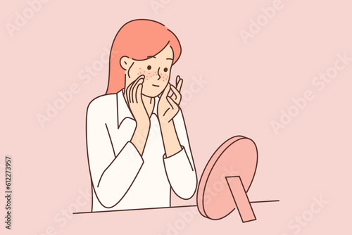 Teenager girl with freckles on face looks in mirror, studying rash or pimples that have appeared