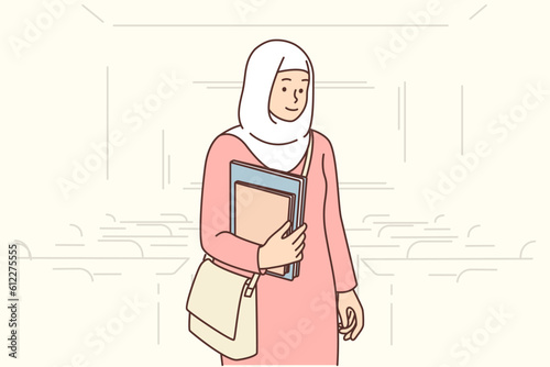 Muslim student girl stands in auditorium with books in hands, dressed in islamic clothing and chador
