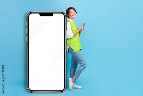 Full size photo of satisfied pretty girl with brown hairdo wear green vest lean on wall hold smart phone isolated on blue color background