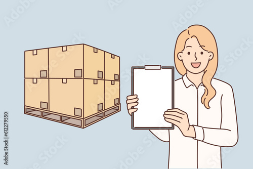 Woman logistician stands near boxes placed on pallet and demonstrates empty clipboard