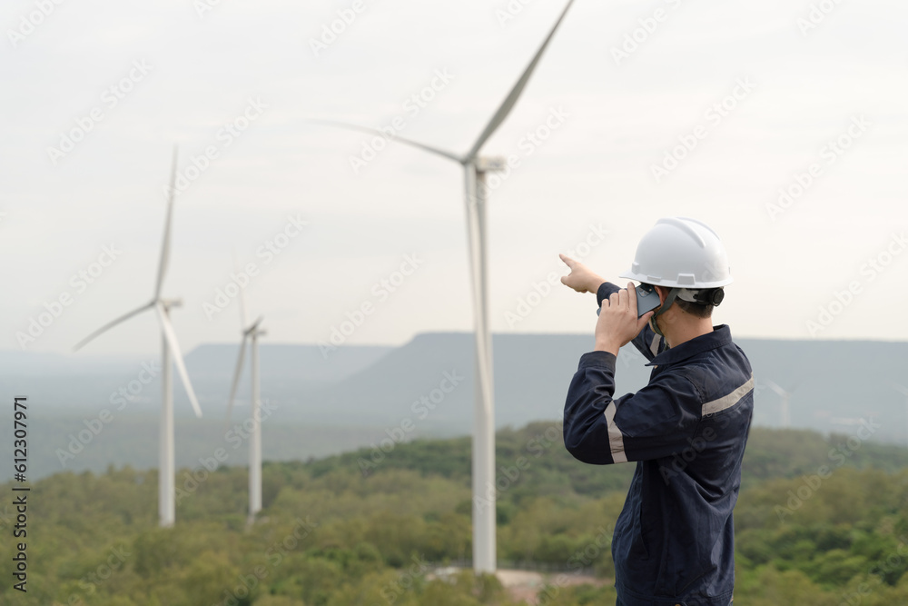 A worker in a wind turbine field looks towards wind turbine while holding a tablet, managing the efficient production of renewable energy.