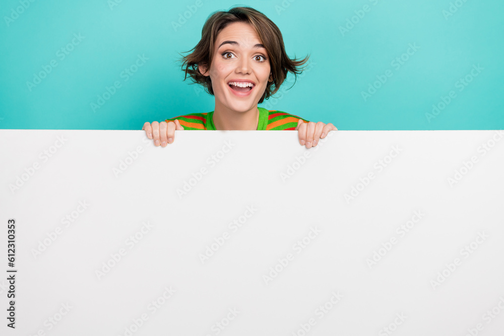 Portrait of positive cute girl toothy smile peeking behind empty space billboard isolated on turquoise color background Portrait of positive cute girl toothy smile peeking behind empty space billboard isolated on turquoise color background