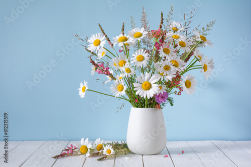 A bouquet of white daisies and other wildflowers in a white vase on a blue background