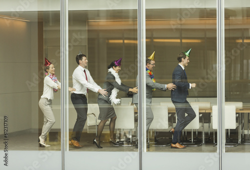 Business people wearing party hats dancing in conga line at conference room window