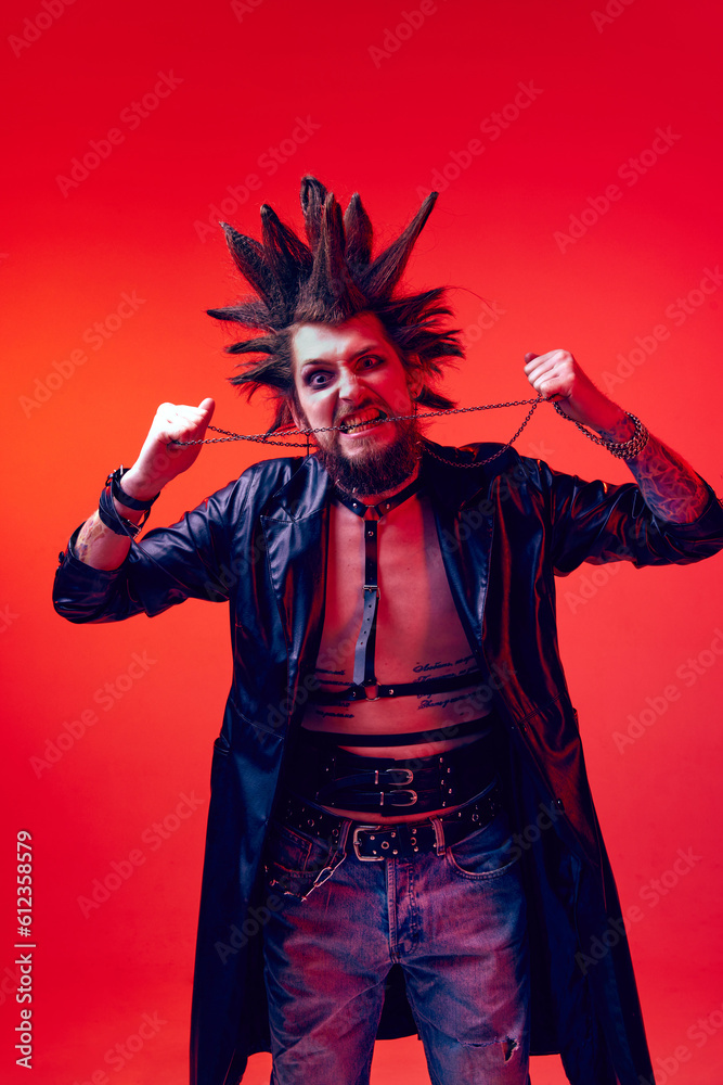 Angry and crazy look. Portrait of young man, punk with crazy hairdo and makeup posing against red studio background in neon light. Concept of music, lifestyle, subculture, art, youth, human emotions Angry and crazy look. Portrait of young man, punk with crazy hairdo and makeup posing against red studio background in neon light. Concept of music, lifestyle, subculture, art, youth, human emotions