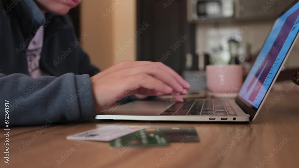 Female hands typing on keyboard laptop using touchpad lying on wooden table next to bank cards, in kitchen. Close up. Work environment concept. Female hands typing on keyboard laptop using touchpad lying on wooden table next to bank cards, in kitchen. Close up. Work environment concept.