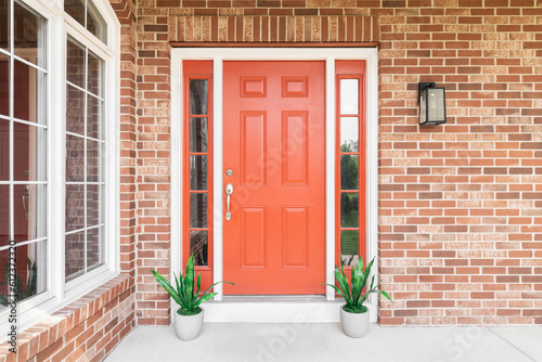 A home's red front door surrounded by red brick and plants sitting in front of the windows.