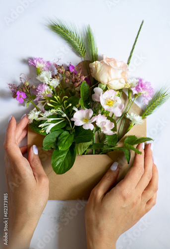Female hands holding an open craft mail envelope full of various flowers on a marble pastel background. Flat lay. Copy space. Minimalistic composition. Concept of love and nature
