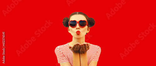 Portrait of stylish young woman blowing her lips with lipstick sending sweet air kiss wearing red heart shaped sunglasses with cool girly hairstyle on background