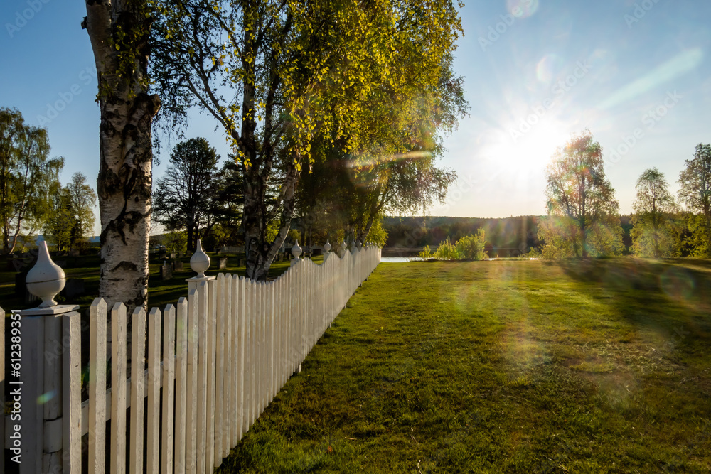 Sorsele, Sweden A white picket fence surrounding the church cemetery in the early morning and a steel arch bridge over the Vindalven river.