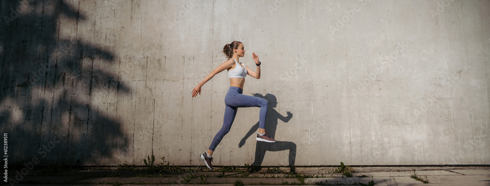 Fototapeta premium Beatutiful female athlete in sportswear exercising in the city in the morning. Fitness woman running in front of concrete wall casting shadow.