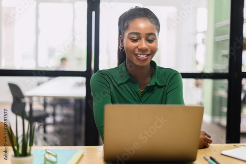 Friendly and positive african-american female employee or student using laptop, multiracial business woman looking at the screen, typing, responding to emails sitting in contemporary office
