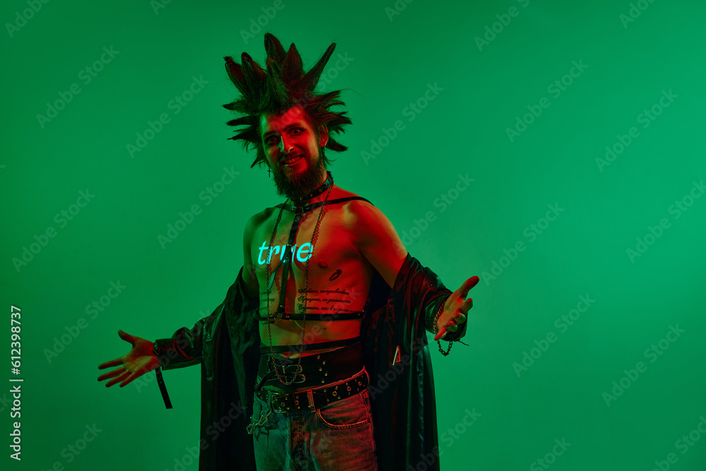 Fototapeta premium Expressive young man, rock musician, punk with neon filter on body posing against green studio background in neon light. True. Concept of music, lifestyle, subculture, art, youth, human emotions