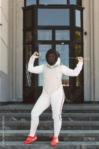 Female Fencer athlete wearing mask and white fencing suit and holding the sword, posing outdoors