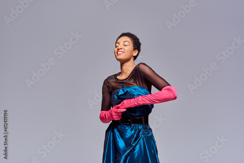 Cheerful and stylish young african american woman in pink gloves and cocktail dress standing with closed eyes isolated on grey, modern generation z fashion concept