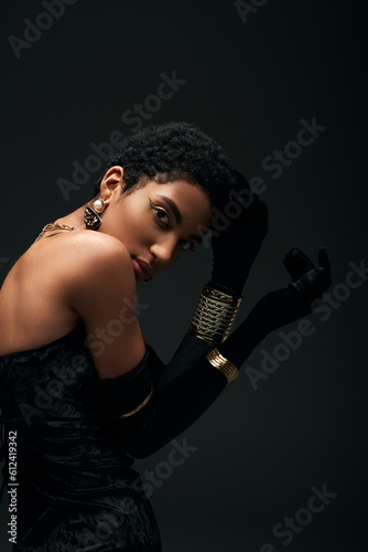 Trendy young african american woman in golden accessories, gloves and dress touching hair and looking at camera isolated on black, high fashion and evening look