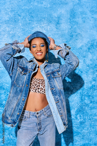 Trendy african american woman with closed eyes and bold makeup touching beret and posing in denim clothes and top on blue textured background, stylish denim attire © LIGHTFIELD STUDIOS