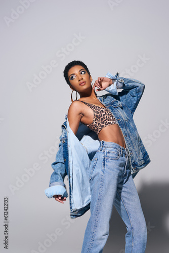 Fashionable african american model with short hair, vibrant makeup and golden earrings wearing denim jacket, top and jeans while standing on grey background, denim fashion concept