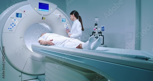 Medical worker adjusts capsule of MRI examination. Patient lies on MRI table and undergoes magnetic resonance imaging. Man in robe and protective cap enters capsule for examination. photo