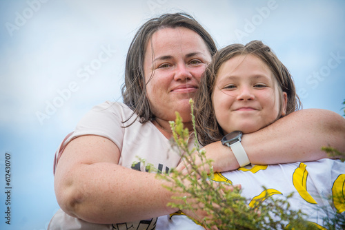 In summer, an overweight mother and daughter stand embracing near the river.