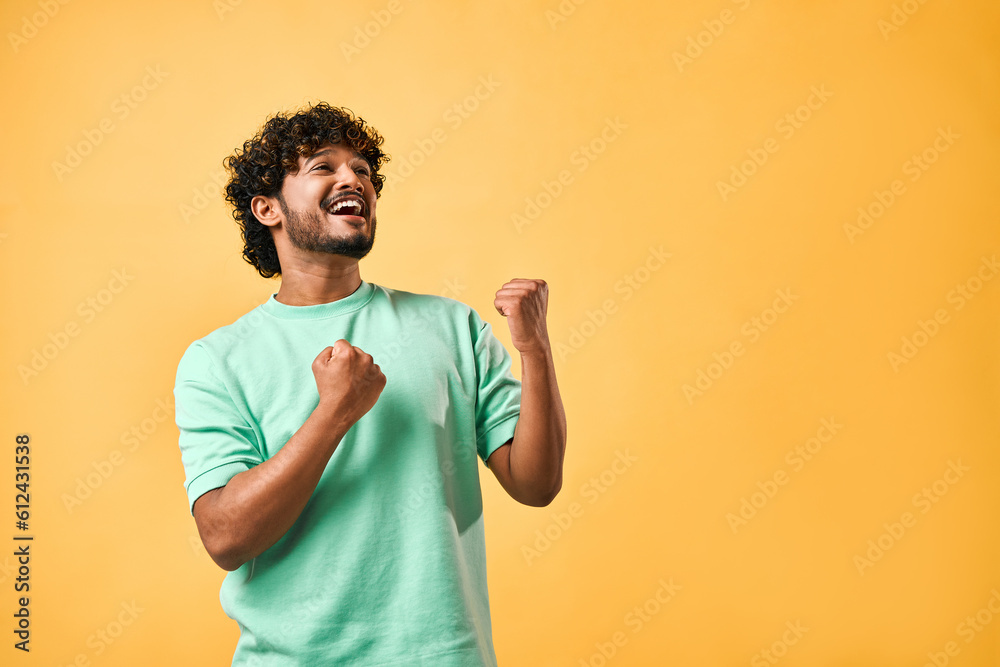 Emotion of joy, celebration of victory. Handsome Indian man is sincerely happy, looking to the side and making a winning gesture while standing against a yellow background. Copy space. Emotion of joy, celebration of victory. Handsome Indian man is sincerely happy, looking to the side and making a winning gesture while standing against a yellow background. Copy space.