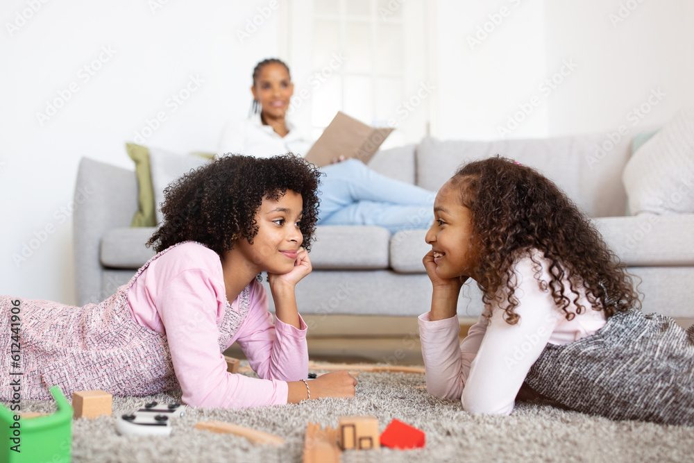 Fototapeta premium Black Sisters Having Fun Playing While Mom Reading Book Indoors