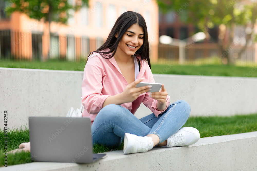 Happy hispanic lady using cellphone playing games and learning online, sitting in college campus or park Happy hispanic lady using cellphone playing games and learning online, sitting in college campus or park