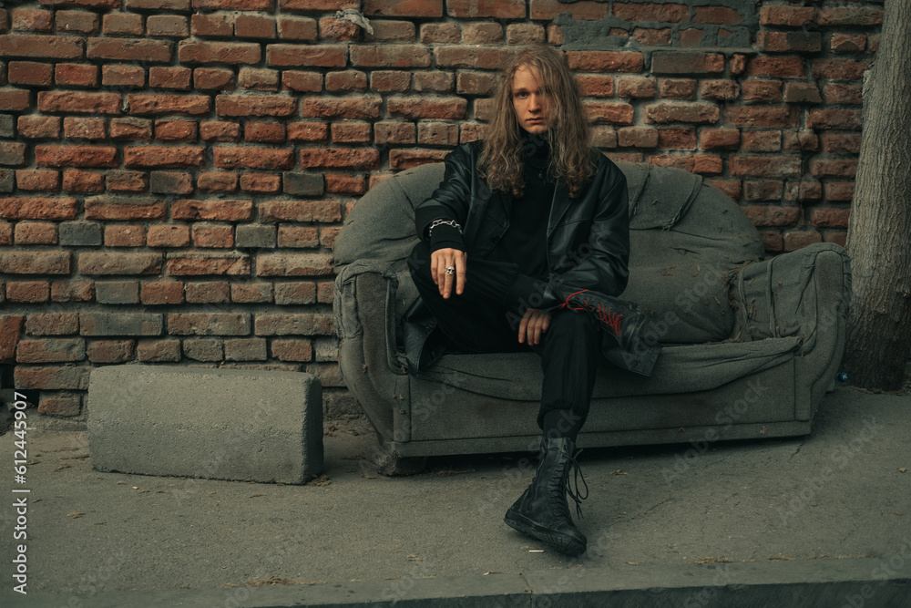 Young male model brutal man wearing black clothes sitting on old sofa on city street Young male model brutal man wearing black clothes sitting on old sofa on city street