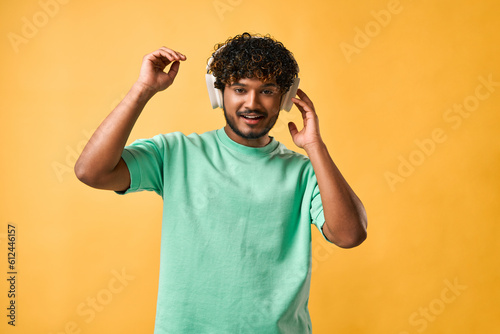 Portrait of a curly-haired Indian man in a turquoise t-shirt adjusting wireless headphones and looking at the camera.
