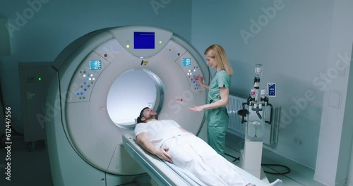 Male patient lies at TC scanner bed. Man waits for examine with MRI capsule. Female doctor dressed up in uniform explained to patient what to do. Medical worker perform magnetic resonance imaging. photo