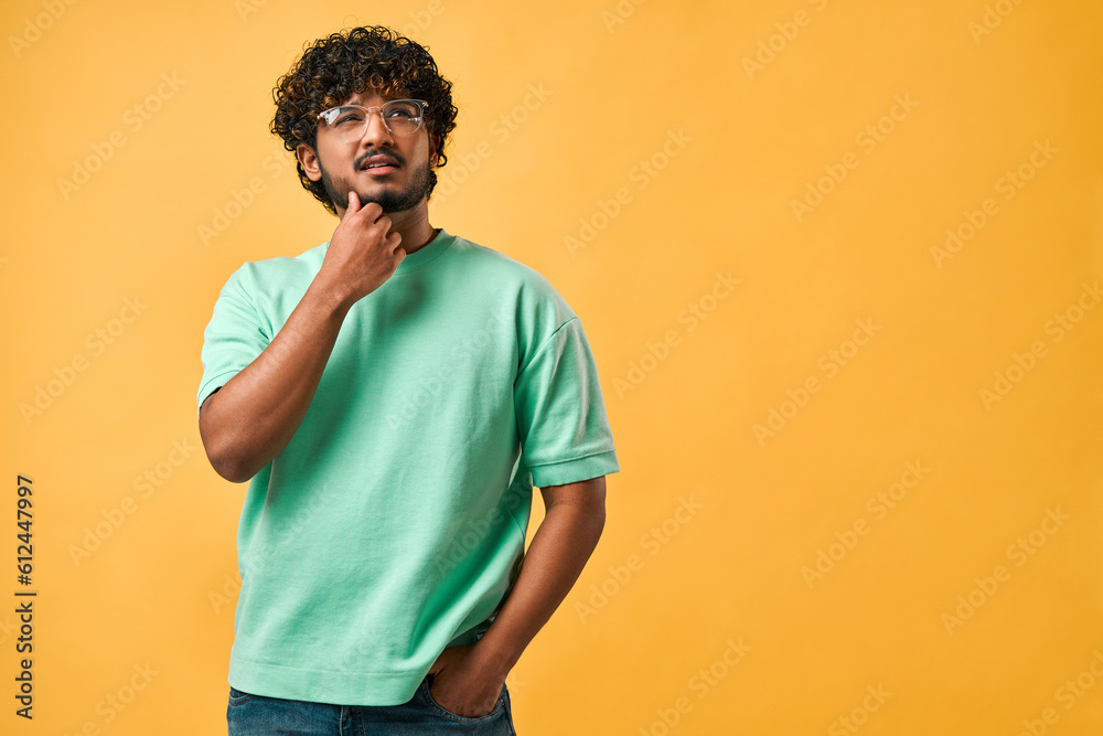 Portrait of a handsome curly-haired Indian man in a turquoise t-shirt and glasses thoughtfully scratching his chin and looking up. The emotion of thoughtfulness, doubt. Copy space. Portrait of a handsome curly-haired Indian man in a turquoise t-shirt and glasses thoughtfully scratching his chin and looking up. The emotion of thoughtfulness, doubt. Copy space.
