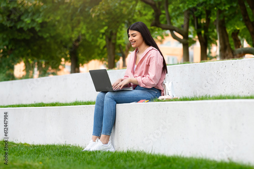 Happy teen hispanic lady with laptop sitting in park, student girl studying remotely with computer outdoors, copy space