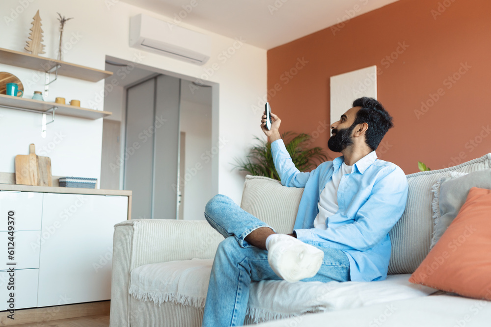 Fototapeta premium Relaxed Indian Man Turning On Air Conditioner Sitting At Home