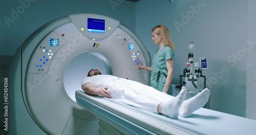 Male patient lies at TC scanner bed. Man goes out of MRI capsule. Female doctor dressed up in uniform have talk with patient. Medical worker gestures and pushes buttons of MRI capsule. photo
