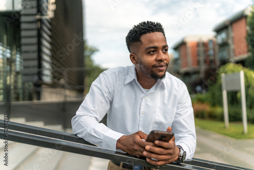 A portrait of a handsome Black businessman leaning against a railing during his break, holding a phone in his hand. With a relaxed posture and a thoughtful expression.